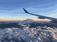 A photograph taken out of the window of a plane. The wing is in view as the sun rises over a snowy capped mountain surrounded by cottony clouds.