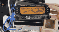 A hand holds a display with knobs and buttons around the perimeter connected with a blue cable to a power supply box. The display is orange and reads REPEATER with an illustration of a satellite in space.