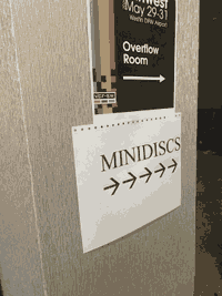 a piece of paper is taped to a column which reads MINIDISCS with five right-facing arrows underneath. it appears haphazardly taped up in contrast to the official event sign above it directing attendees to the panel overflow room.
