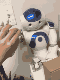 A blue and white humanoid robot (Aldebaran NAO) leaning against a wall. A human hand is manipulating its fingers.