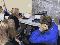 3 people sitting around an unfolded table, there are snacks around and there are two laptops at the table displaying a powerpoint presentation with space mission logos and Inkscape with an orbital trajectory image being made respectively.