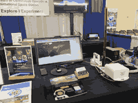 A picture of objects on a table with a black tablecloth. A monitor displays an orbital display and there is an antenna made out of a tape measure that extends in a t-shape in front of a white box.