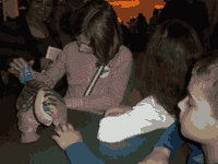 Three children crowd around a robotic dinosaur. A woman is smiling in the background.